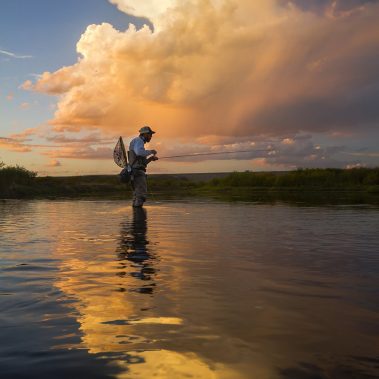 A fly fisherman standing in a river at sunset.