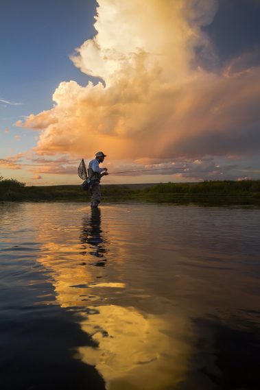 A fly fisherman standing in a western river with a sunset in the back ground