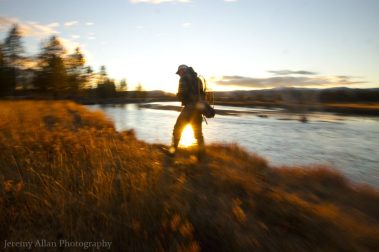 A fly fisherman walking a river bank at last light