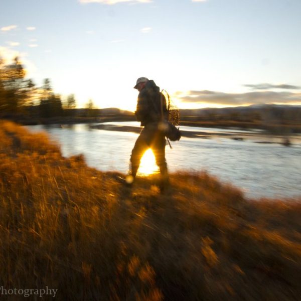 A fly fisherman walking a river bank at last light