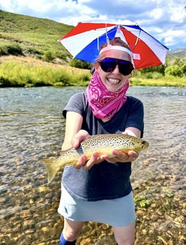 A happy fly fisher lady holding a brown trout while wearing an umbrella hat.