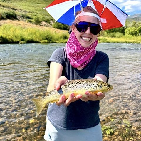 A happy fly fisher lady holding a brown trout while wearing an umbrella hat.